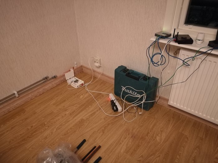 Rördragning i en hörna av ett rum med verktyg och material på golvet och oordnade kablar hängande från en väggmonterad router.
