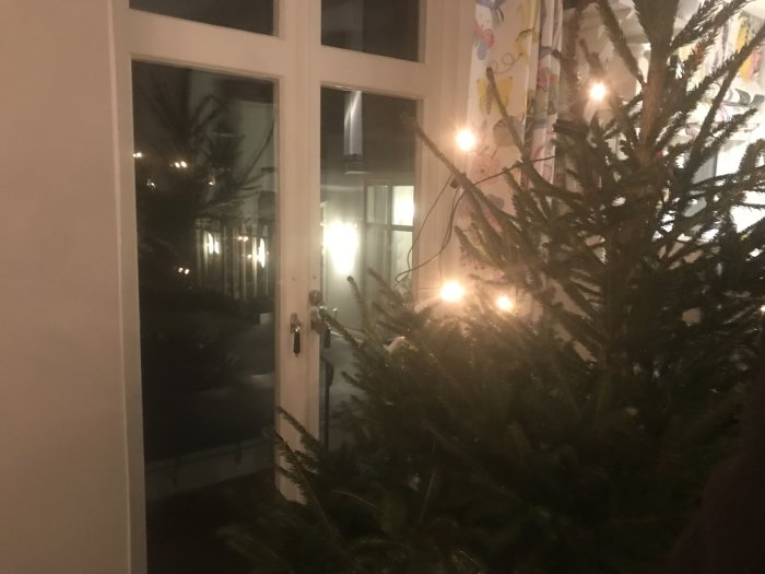 Oupplyst julgran nära ett fönster inomhus på kvällen, med svagt ljus från utsidan.