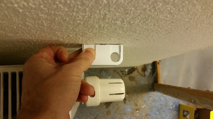 Hand som håller en termostat under en takyta där rörinstallation diskuteras, med lite plats till väggen.
