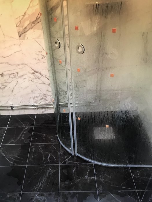Nyligen renoverat badrum med en duschkabin där vatten rinner ut mot dörren på svarta plattgolv.