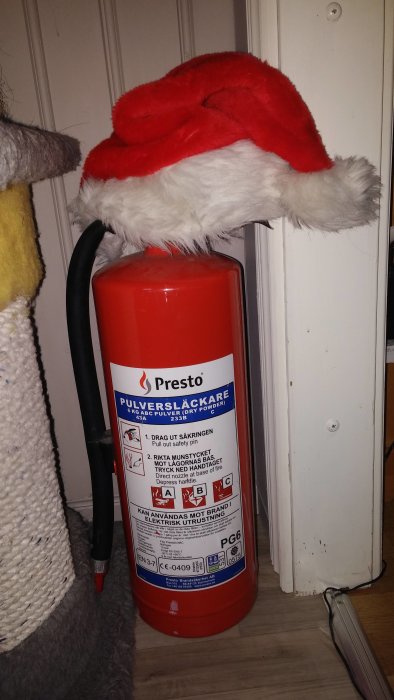 Brandsläckare med en julig tomteluva på toppen mot vit dörrbakgrund.