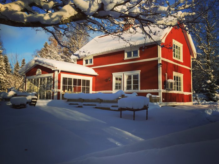 Röd trävilla med snötäckt mark och trädgårdsmöbler, omgiven av snöklädda träd och solljus.