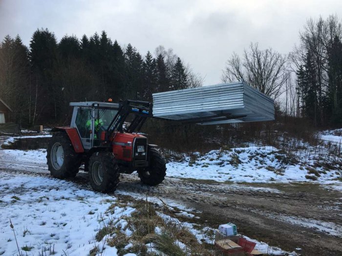 Traktor med lastade stallpaneler på långgafflar i snöigt landskap.