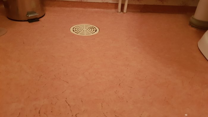 Rosa plastmatta på golv i badrum med en central golvbrunn och toalettartiklar i bakgrunden.
