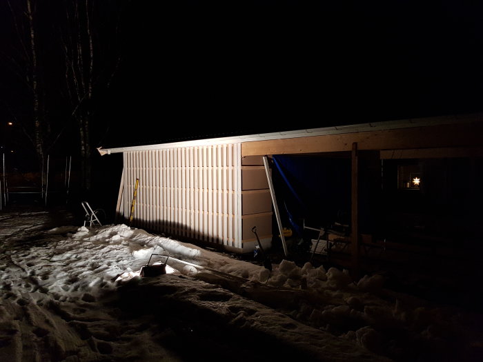 Nypanelad väggsida av ett hus i mörker med artificiellt ljus och snö på marken.