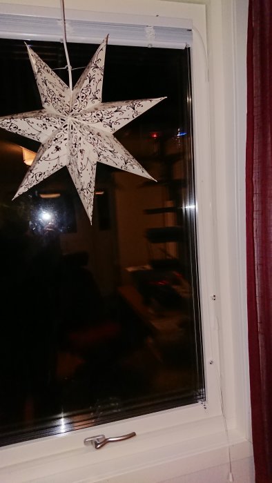 Dekorativ pappersstjärna hängande framför ett fönster med persiennmontering och synliga snören vid fönsterkarmens botten.