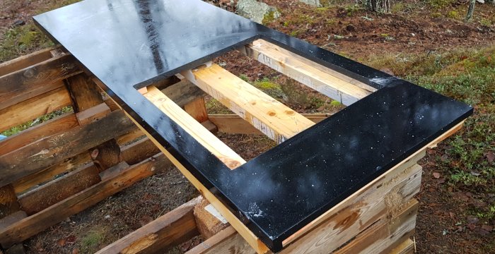 Svart bänkskiva med utskurna hörn placerad på träbockar utomhus, synligt damm och skärspån.