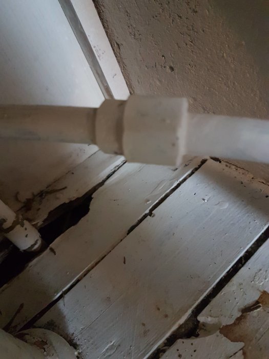 Vit målad klämringskoppling för vattenrör framför vitmålade golvbrädor.
