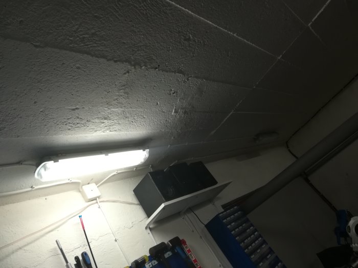 En tänd lysrörsarmatur på ett garage tak med släckta lampor i bakgrunden, verktyg på hyllor nedanför.