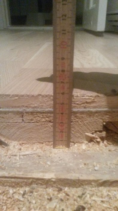 Mätsticka visar tjockleken på ett golvunderlag i en renoveringsprocess, med synliga lager av golvspån och OSB.
