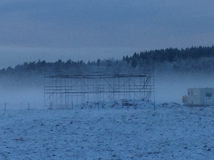 Byggställning på en husgrund i ett snötäckt landskap med dimma i bakgrunden.