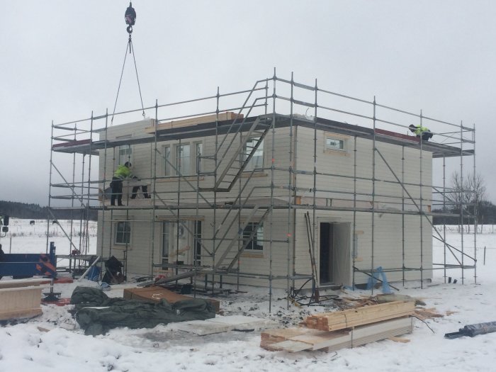 Två arbetare bygger ett tvåvåningshus med ställningar runtom i vinterlandskap.