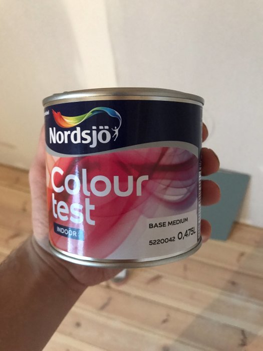 Hand som håller en Nordsjö Colour Test-färgburk med bakgrund av en målad vägg i mörk färg.