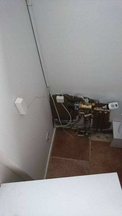 Värmefördelare och rör vid en hörnvägg med synlig kabeldragning och termostat.
