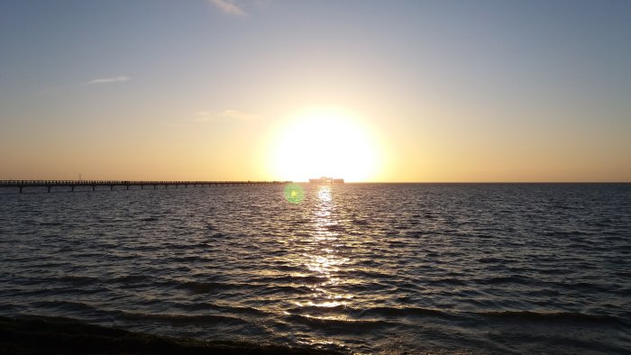 Solnedgång över havet med en lång pir och ett hus på vattnet, reflektioner i vattenytan.