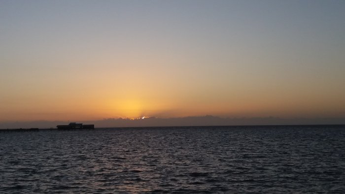 Solnedgång över havet med silhuetten av en pir och byggnader mot den ljusa himlen.