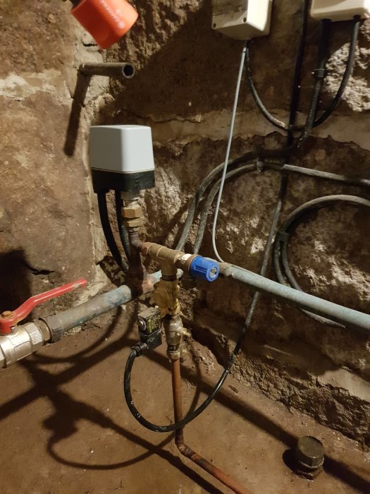 Källarkomponenter med termostatstyrd magnetventil, rör och elektriska anslutningar på en rå stenvägg.