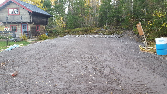 Grusad uppfart med uppradade betongplintar framför ett timmerhus, stödmur av sten och skog i bakgrunden.