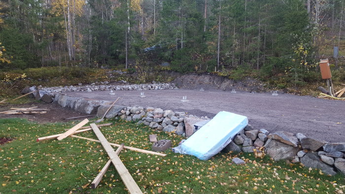 Byggplats med grusad mark, stödmur av natursten, plintar som sticker upp, och byggmaterial på marken.