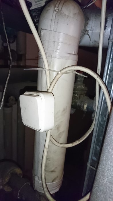 Vit avloppsrörinstallation med kabeldragning och en strömbrytare fäst på ett rör, i en mörk och odefinierad miljö.
