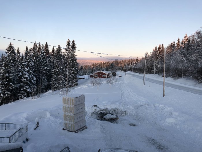 Vinterlandskap med snötäckt mark, träd och hus i bakgrunden, pall med material i förgrunden.