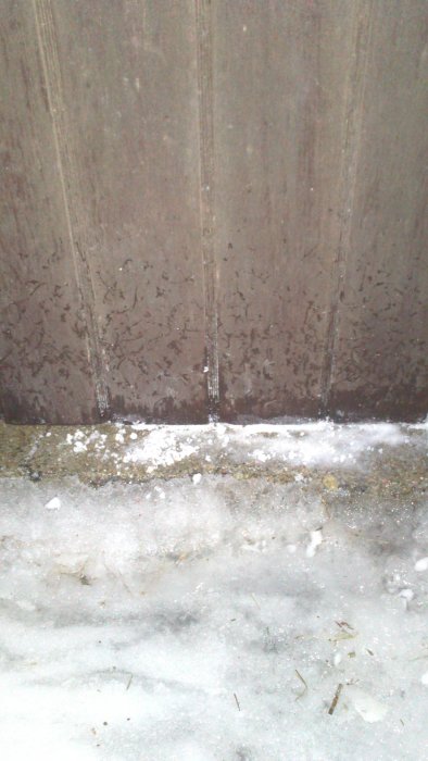 Mörk träport med synliga regndroppar, möter ett frusset snötäcke vid tröskeln.