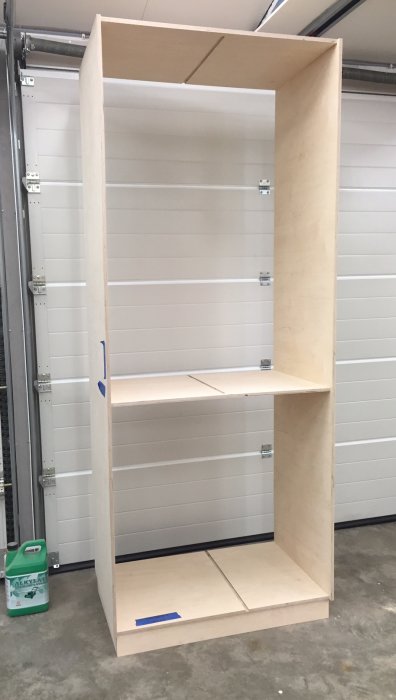 Plywoodskåp under konstruktion i ett garage med synliga stödfästen och ojämna hyllplan.