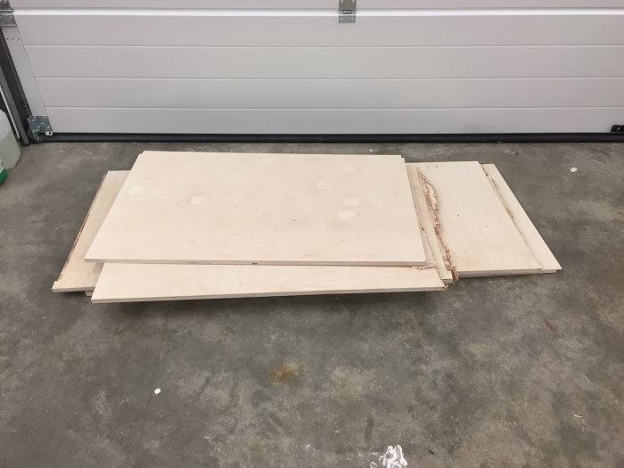 Oanvända plywoodskivor ligger på ett garagegolv, några skivor ser skadade ut.