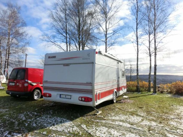 Röd skåpbil och vit husvagn på en gräsmatta med snöfläckar, träd och himmel i bakgrunden.