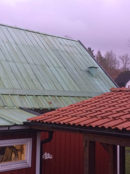 Ett rött hus med ett grönt plåttak och ett tegeltak i förgrunden, ventilationstrumma synlig på plåttaket.