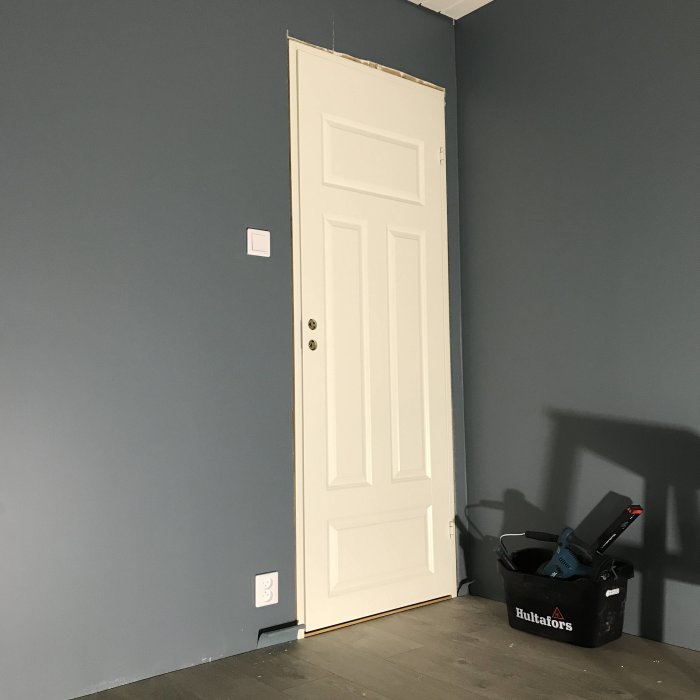Vit panel dörr och dörrkarm installerade i en mörkblå vägg, renoveringsverktyg syns i förgrunden.