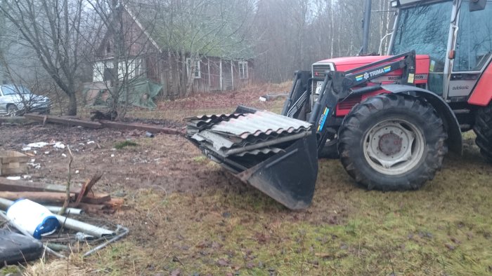 Traktor med frontlastare håller eternittakplattor, omgiven av byggavfall och nedfallna grenar.