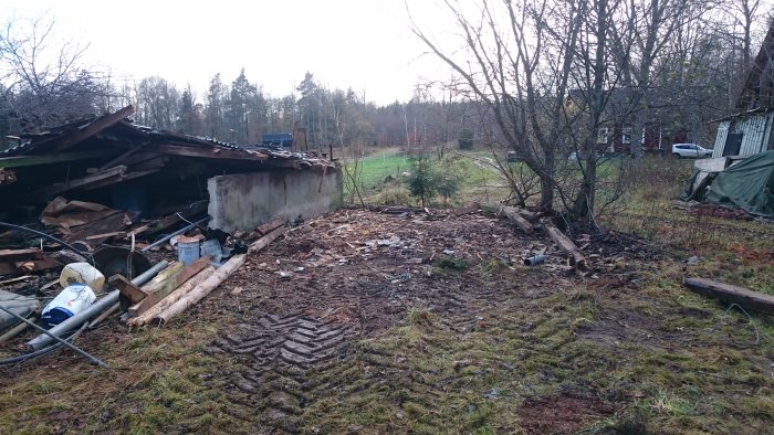 Raserat stall med spridda byggrester och skrot på en övergiven tomt med träd i bakgrunden.