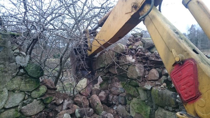 Gul grävmaskin från JCB river stenmur bland vintergrenar.