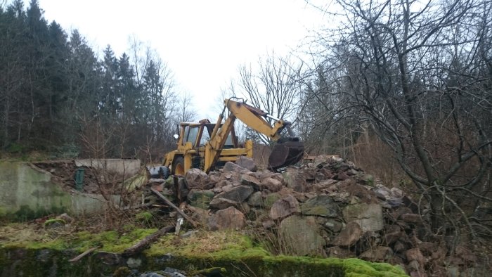 Gul grävmaskin hanterar skrot och stenar vid en raserad byggnad, omgiven av träd och grönska.