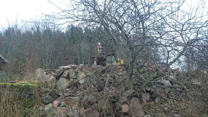 Gul grävmaskin synlig bakom hög av sten och grenar på en byggarbetsplats.