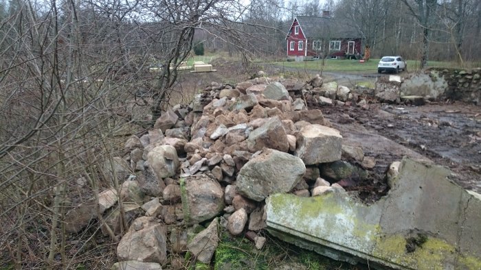 Hög med borttagna stenar och raserade murar vid en renoverad byggnad, med hus och bil i bakgrunden.