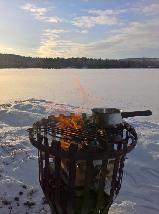 Grill över öppen eld på snötäckt mark med en kastrull på gallret, skog och frusen sjö i bakgrunden.