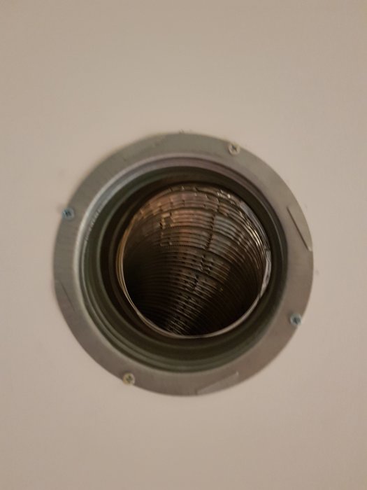 Ventilationsrörskoppling fastskruvad mot en vit vägg, synliga skruvhål på kopplingens fläns.