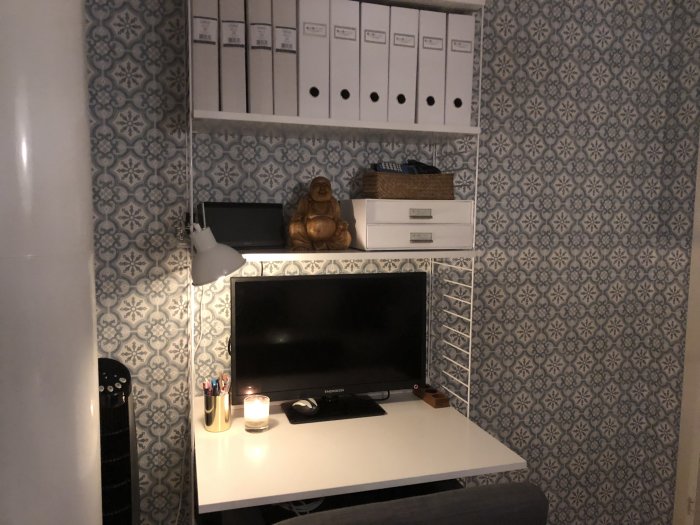 Hörn av ett rum med skrivbord, dator, stringhylla, vita förvaringslådor och Ikea Maskroslampa, mot mönstrad tapet.