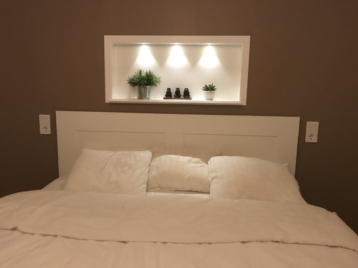 Nyinredd sovrumsinteriör med en vit säng och ovanliggande nisch med belysning samt dekorativa föremål.
