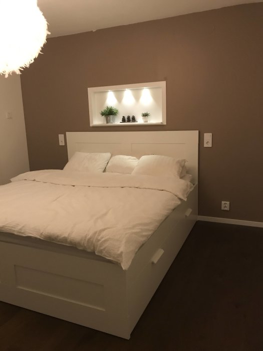 Nyrenoverat sovrum med en vit säng, beige väggar och inbyggd vägghylla med belysning.