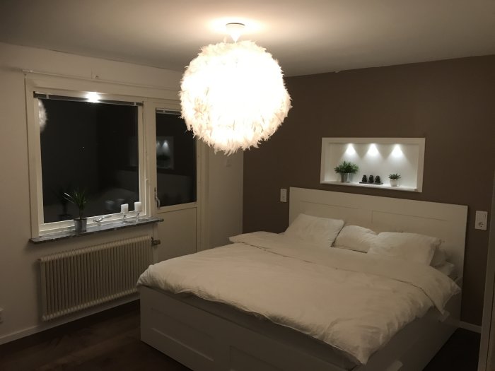 Nyrenoverat sovrum med upplyst säng, taklampa, nisch med belysning och fönster utan gardiner.