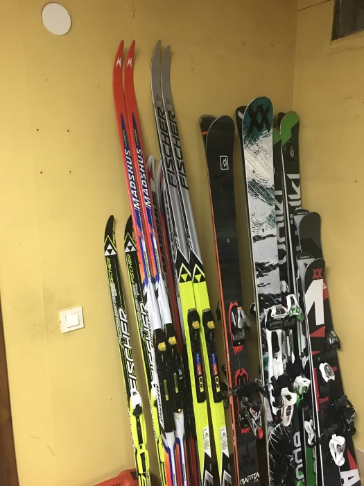 En samling av skidor och snowboards står lutade mot en gul vägg intill varandra.