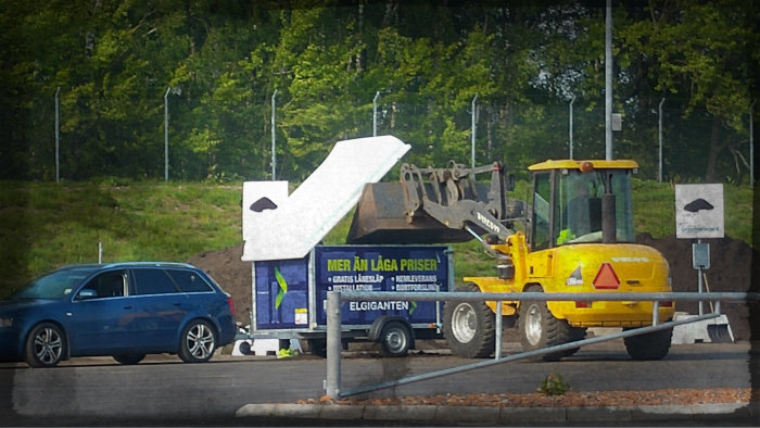 Gul grävmaskin lägger skopa grus på blå bilsläp med Elgiganten-reklam på återvinningsgård.