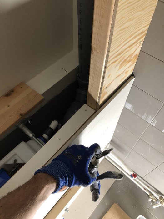 Person i arbetskläder spänner en träregel mot en plywoodskiva i ett pågående byggprojekt, med rörinstallationer i bakgrunden.