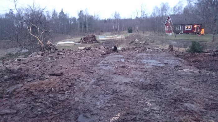 Schaktat område med stenar och jord, delvis rensad platta, i bakgrunden en hög med sten och ett rött hus med julbelysning.