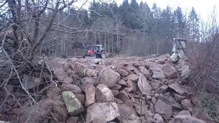 En traktor nära en hög av bortschaktade stenbumlingar bland träd och buskar.