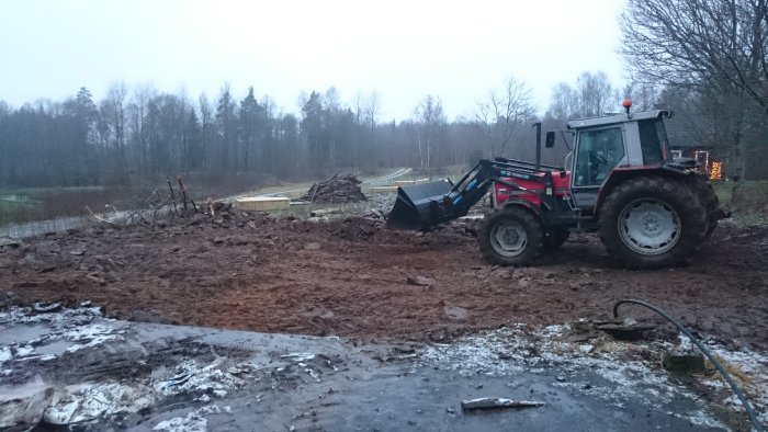 Traktor med skopa som arbetar med att schakta bort sten och jord vid rivningen av ett stall.