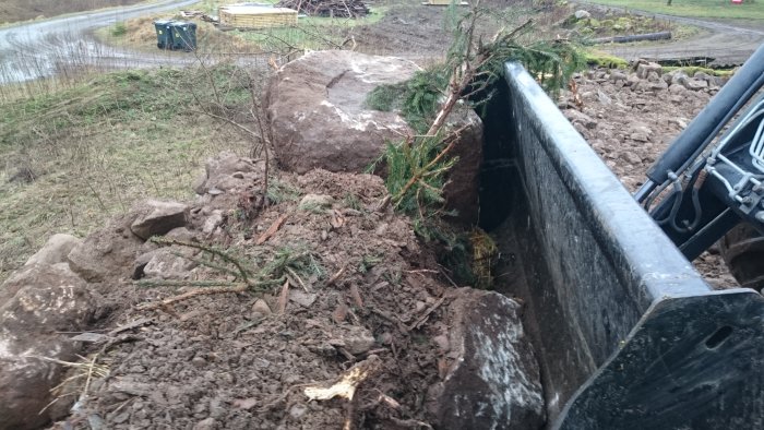 Grävmaskinsskopa fylld med jord och stora stenbumlingar vid arbete på tidigare stallplats.